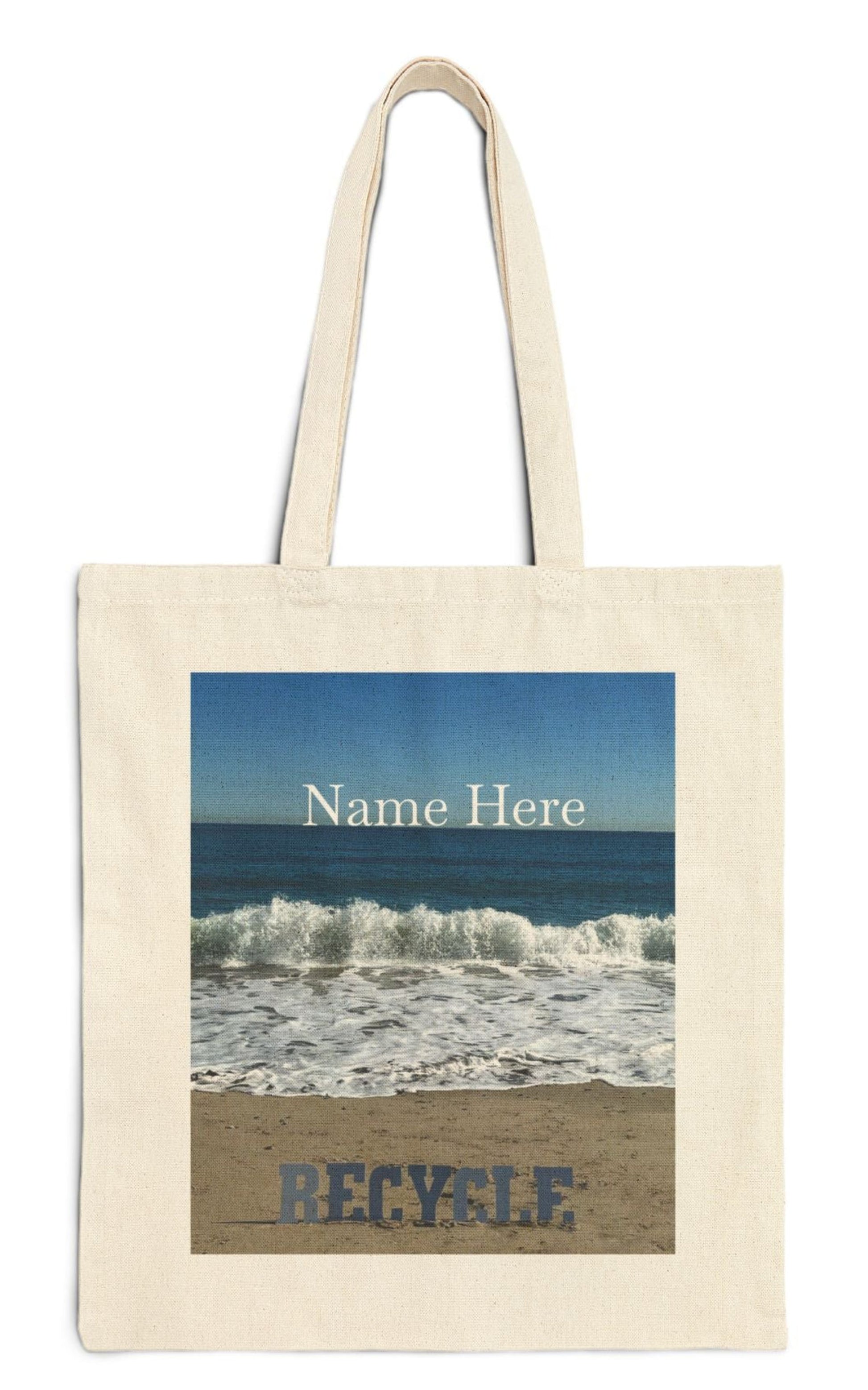 personalized recycle tote bag in natural color the recycle letters are on a sandy shore backed by waves from a blue sea with a blue sky backdrop.